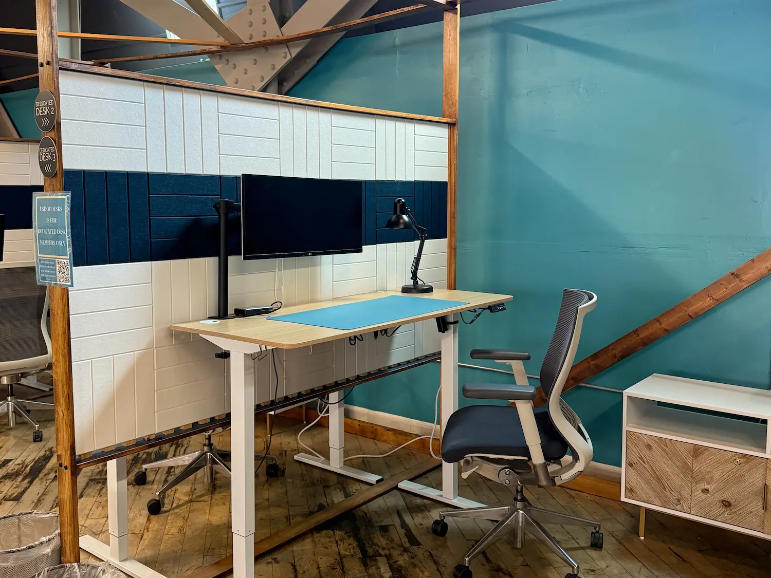 Modern office workspace with an adjustable desk, ergonomic chair, dual monitors, and desk lamp. Teal wall with white panel accents adds a fresh feel.