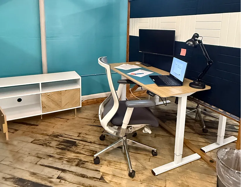 Modern home office with standing desk, ergonomic chair, and laptop against a teal and navy acoustic wall. Bright, even studio lighting on wood floors.