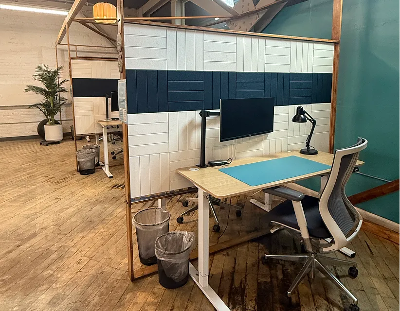 Modern office coworking space with individual desks, white and navy acoustic privacy panels, ergonomic chairs, and warm lighting on hardwood floors.