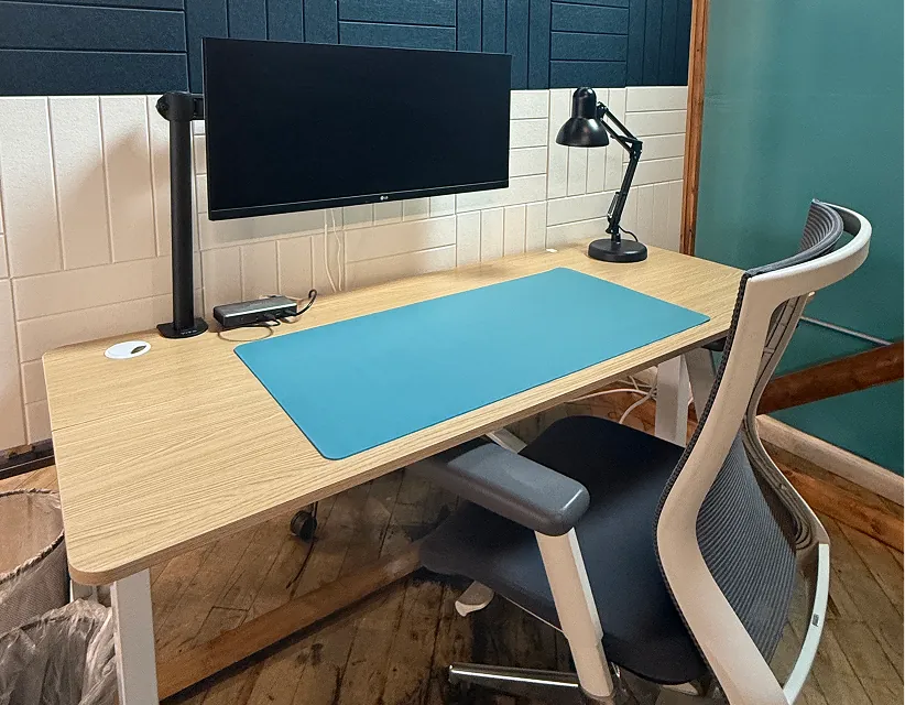 Minimalist home office setup with LG monitor, wooden desk, teal mat, and ergonomic chair against acoustic panel wall, brightly lit by a desk lamp.