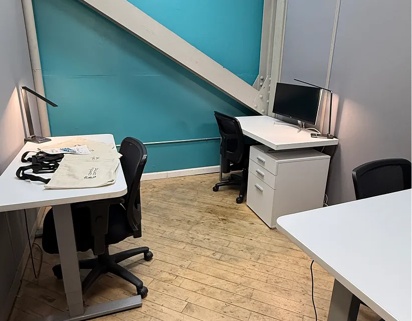 Modern open-plan office with white desks, ergonomic chairs, and potted plants under blue pendant lighting. Industrial ceiling and bright, clean workspace.