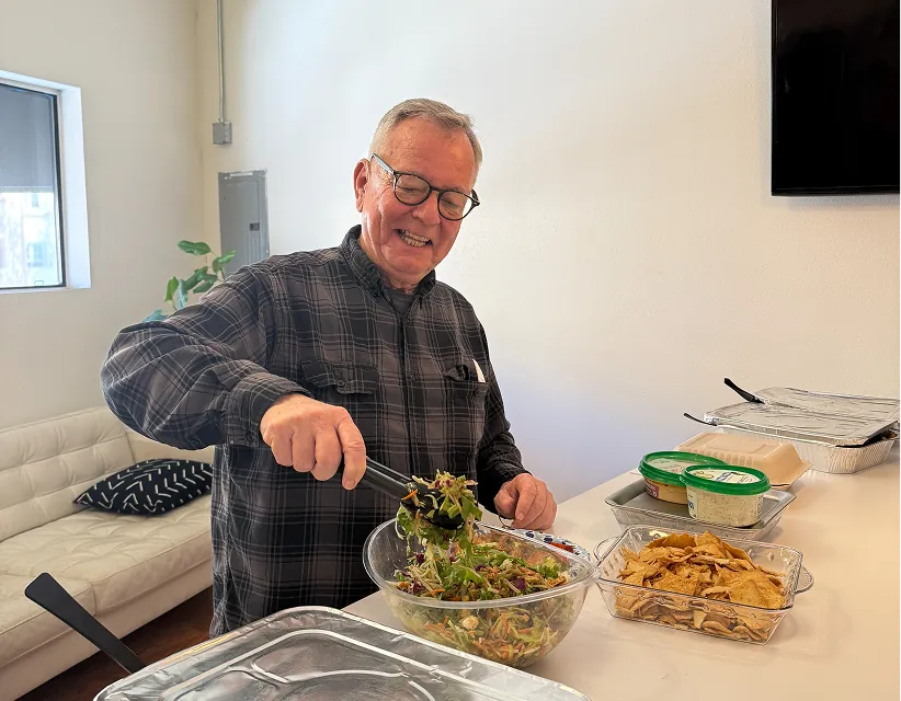 Smiling man in plaid shirt serving salad from bowl at a brightly lit office catered lunch, featuring Sabra branding on dips and pita chips.