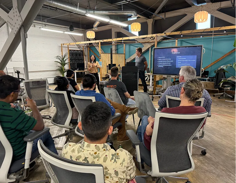 Speaker leads a workshop in a bright loft office, Hapnyn branding on a shirt, with attendees focused on an exercise displayed on a monitor.