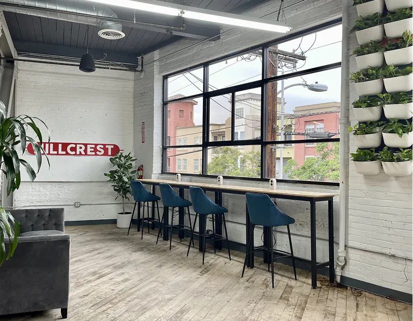 Modern office space with Hillcrest signage, blue bar stools at a window-facing desk, lush vertical wall planters, and bright industrial lighting.