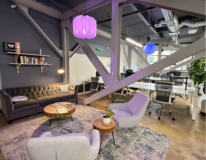 Modern coworking space with industrial steel beams, ambient purple pendant lighting, grey tufted sofa, and "I Love Hillcrest" sign in a bright office.