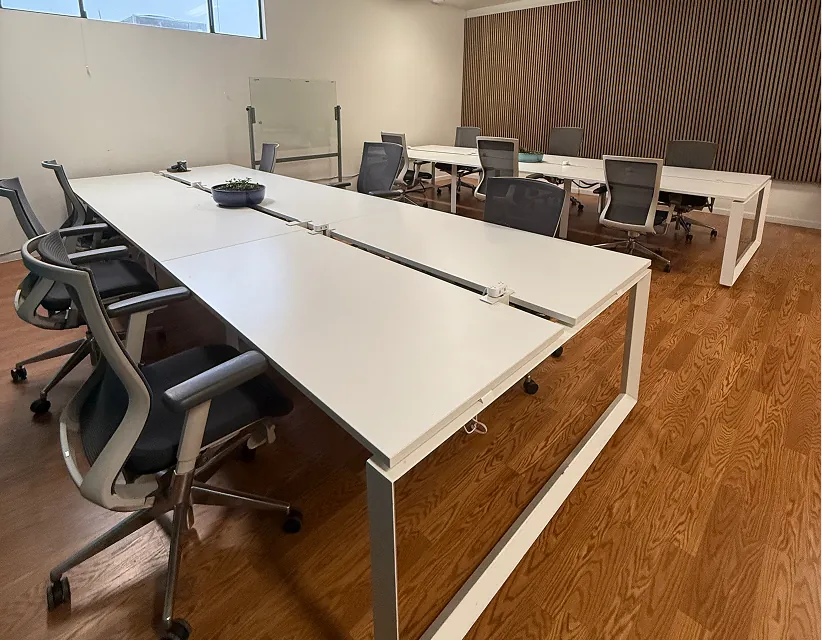Modern office workspace with white desks, ergonomic chairs, and wood flooring, illuminated by soft natural light with wooden slat wall accents.