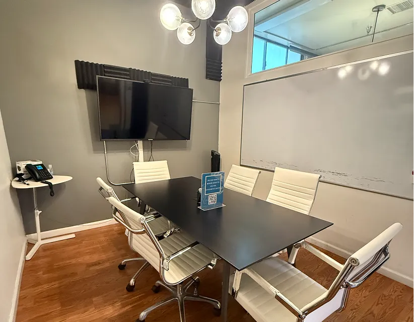 Modern meeting room with wood floors, black table, and white chairs. Wall-mounted TV, whiteboard, and reservation sign under warm overhead lighting.