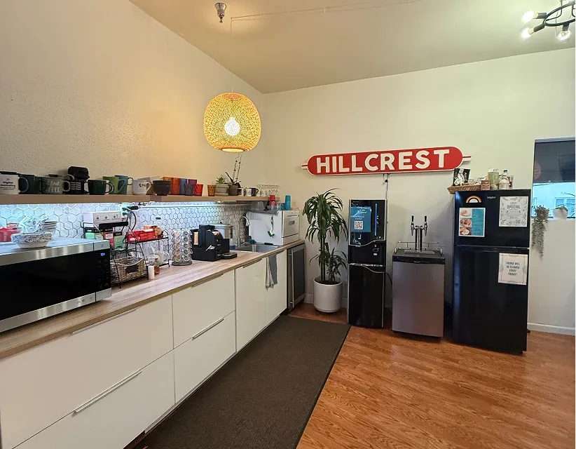 Bright office breakroom with white cabinetry, wood floors, a "HILLCREST" wall sign, coffee station, water cooler, and warm pendant lighting.
