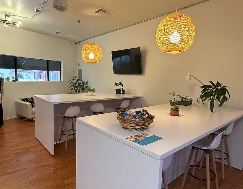 Modern shared office breakroom featuring two white high-top tables, rattan pendant lighting, snacks, and lush green plants under warm ambient light.