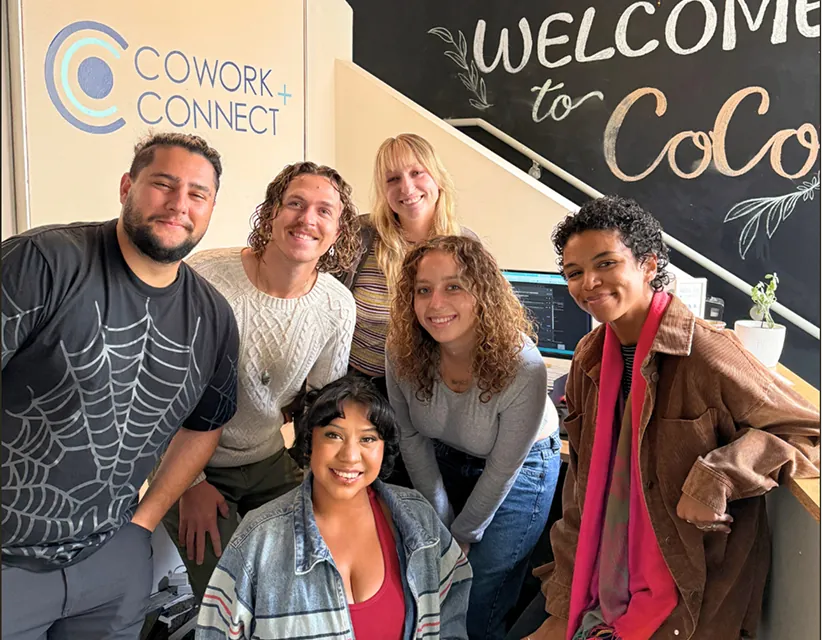 Diverse team smiling under soft lighting in a Cowork+Connect office, featuring branded wall signage and a "Welcome to CoCo" chalkboard background.