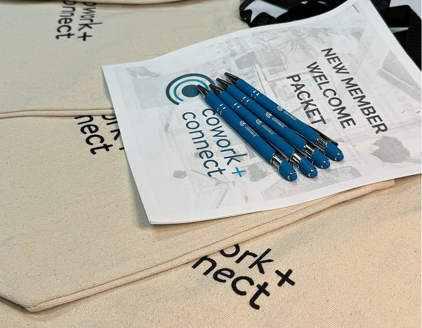 Cowork+Connect branded welcome packet and blue pens on canvas tote bags in bright, even studio lighting.