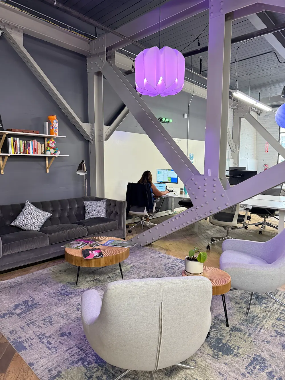 Modern industrial office lounge with a purple pendant light, grey velvet sofa, accent chairs, and an team member working in the background.